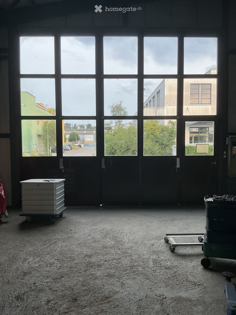Empty warehouse space with large windows, concrete floor, and minimal equipment like a cart and a barrel.