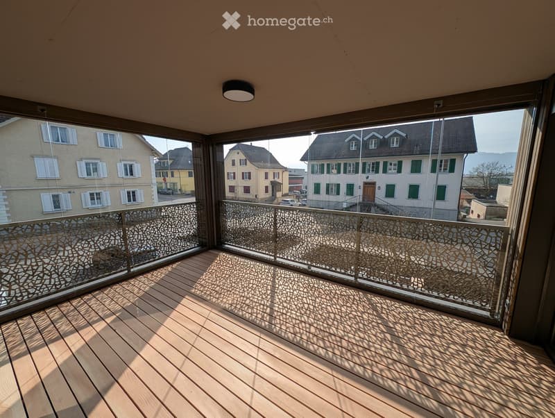 Balcony with wooden decking, glass railing, and decorative metal balustrade pattern. Offers a view of neighboring houses and a clear sky.