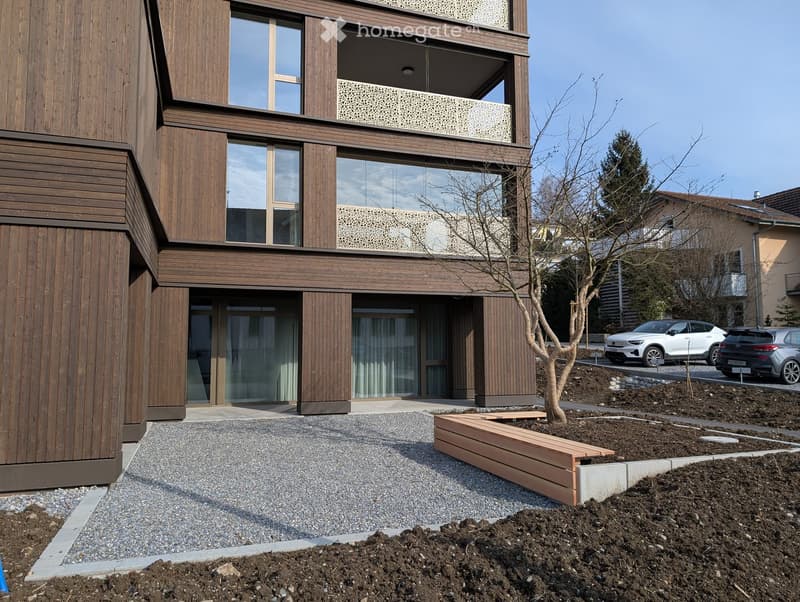 Modern two-story building with wooden facade, large windows, and a balcony. The ground floor has a paved entrance area with a small garden bed and a young tree.
