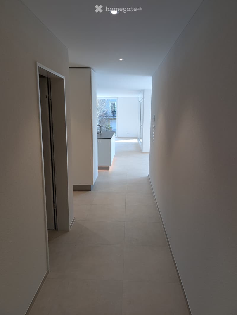 Modern hallway with tiled floor, recessed ceiling lights, and a visible kitchen area with white cabinets and a sink.