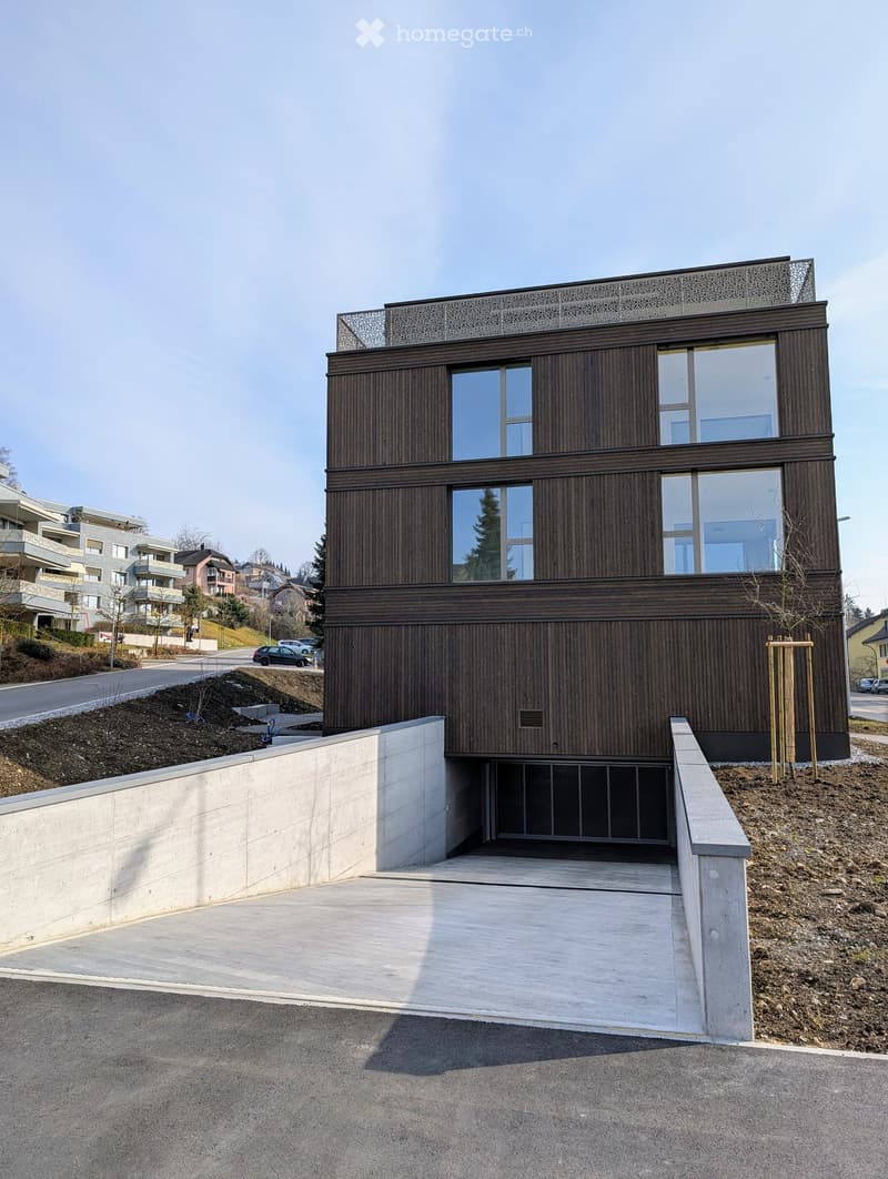 Modern two-story building with dark wooden facade and large windows, featuring a raised ground floor and a concrete entrance ramp.