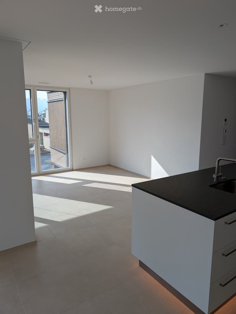 Empty kitchen with tiled floor, white walls, and a black countertop with a sink. Sunlight streams in through a large sliding glass door leading to a balcony.