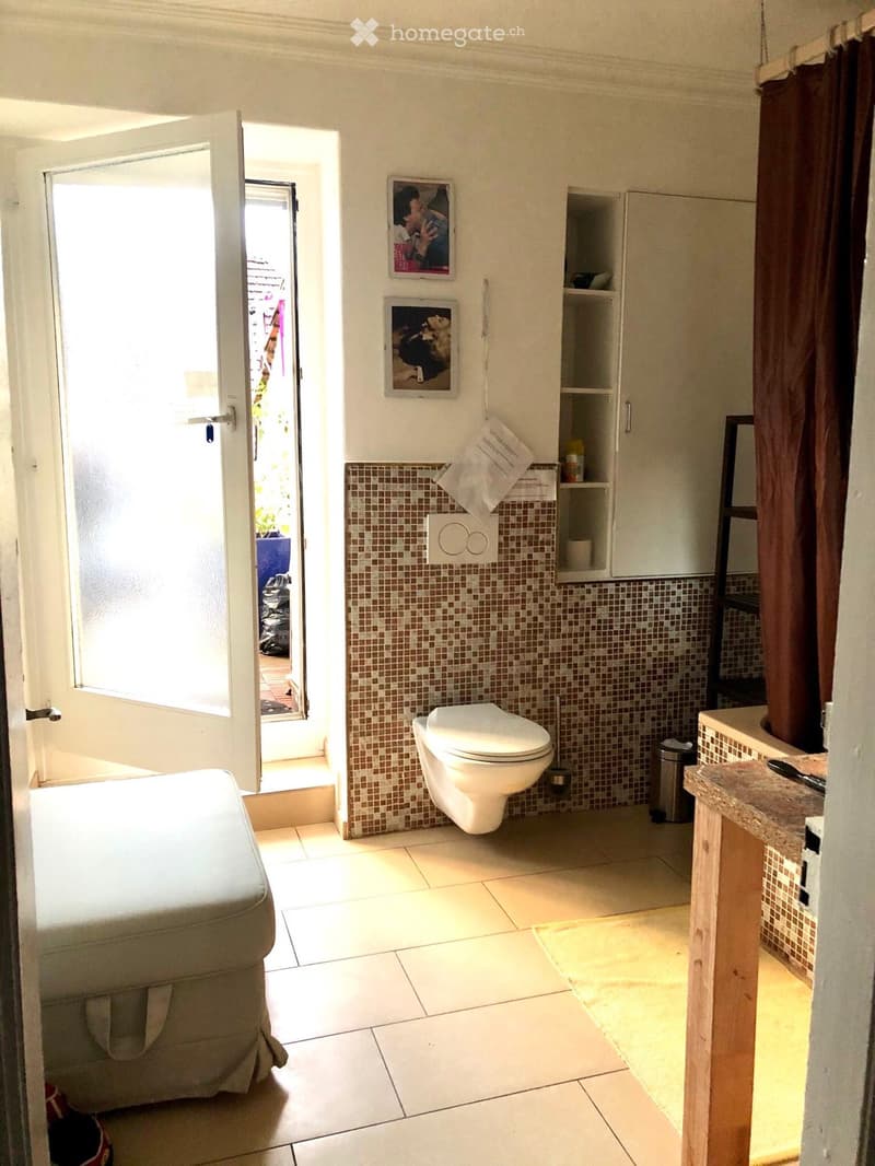 Bathroom with white tiled floor, mosaic tile wall, toilet, sink, cabinet, and picture frames on the wall. An open door leads to an outdoor area.