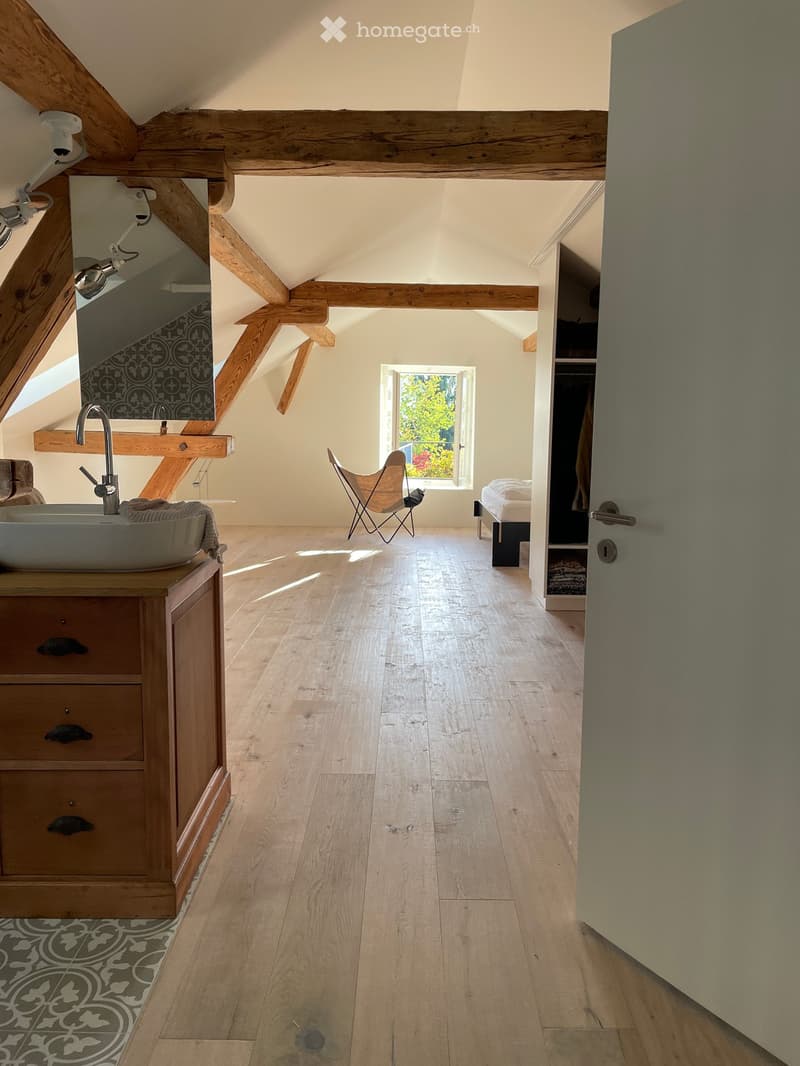 Bright, airy attic space with wooden beams, light wooden flooring, a sink area with a mirror, a chair, and a bed visible in the background.