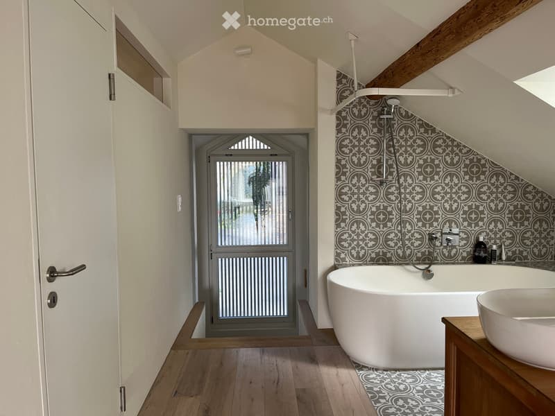 Modern bathroom with a freestanding bathtub, sink, and shower area. The walls feature decorative tiles, and the floor is wooden.