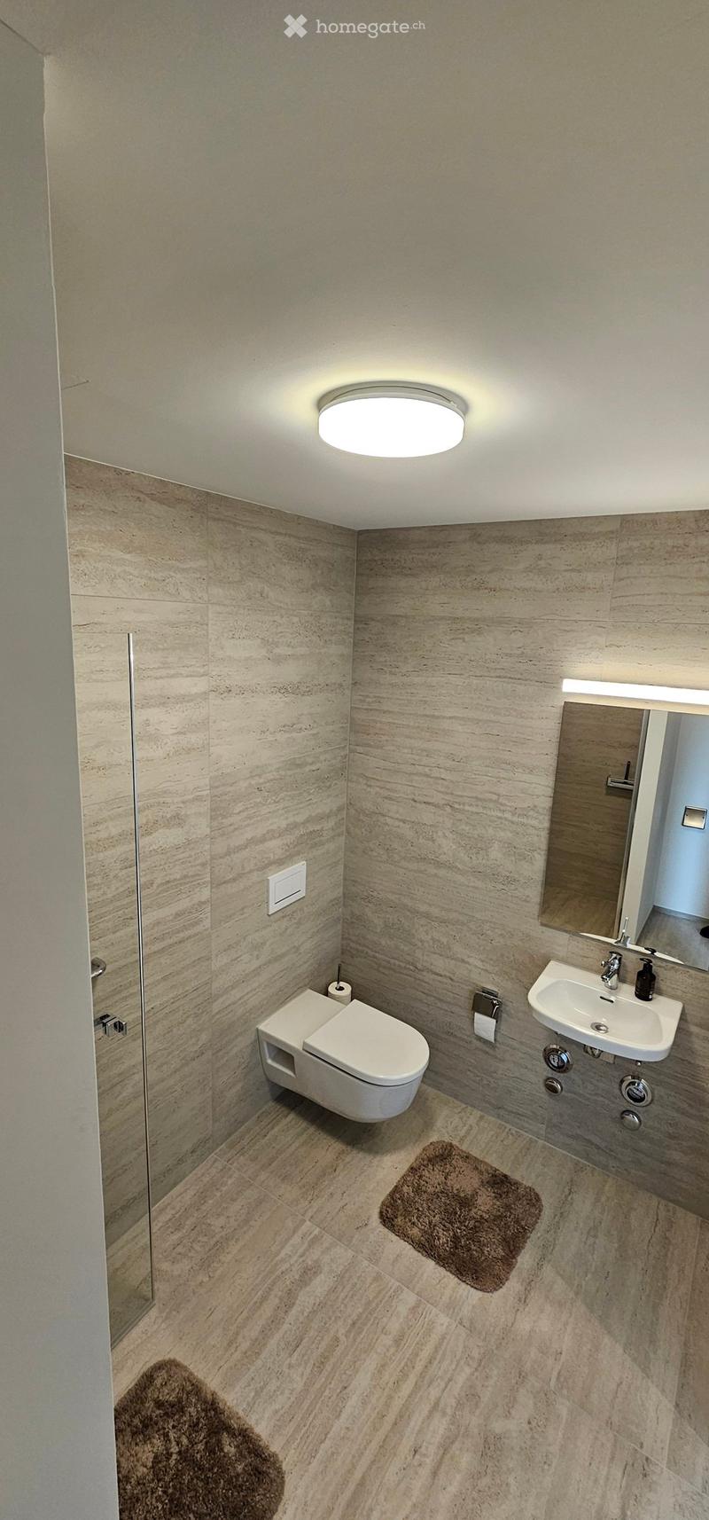 Modern bathroom with marble walls and floor, toilet, sink, mirror, and a round ceiling light.
