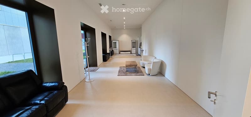 Modern, minimalist living room with white walls, light-colored flooring, and contemporary furniture including a black leather sofa, white chairs, a coffee table, and a coat rack.