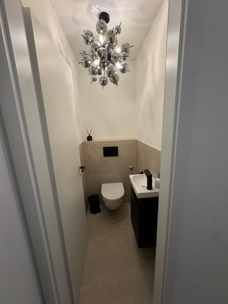 Modern, compact bathroom with a toilet, sink, and a stylish light fixture. Beige tiles and white walls.