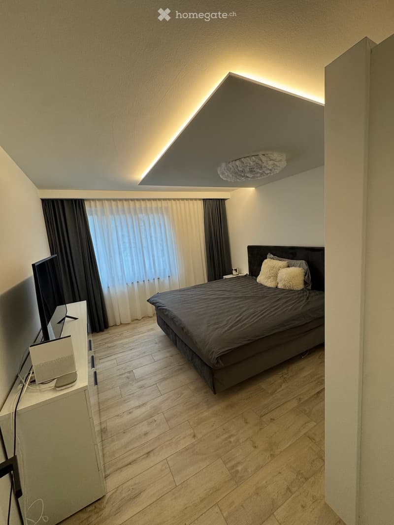 Modern bedroom with a large bed, nightstand, and a TV screen on a white cabinet. The room features wooden flooring, large windows with curtains, and a stylish ceiling light fixture.