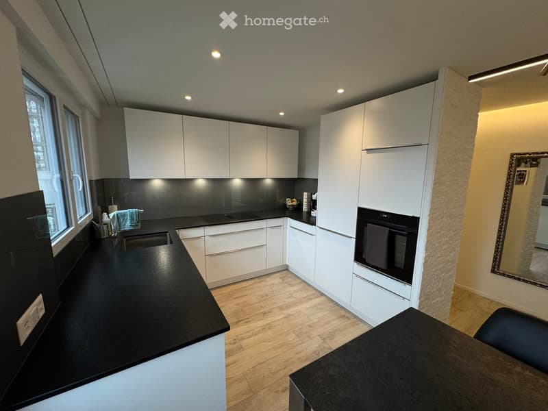 Modern kitchen with white cabinets, black countertops, and a built-in oven. Features a small dining area with a black table and chair, and a large window letting in natural light.