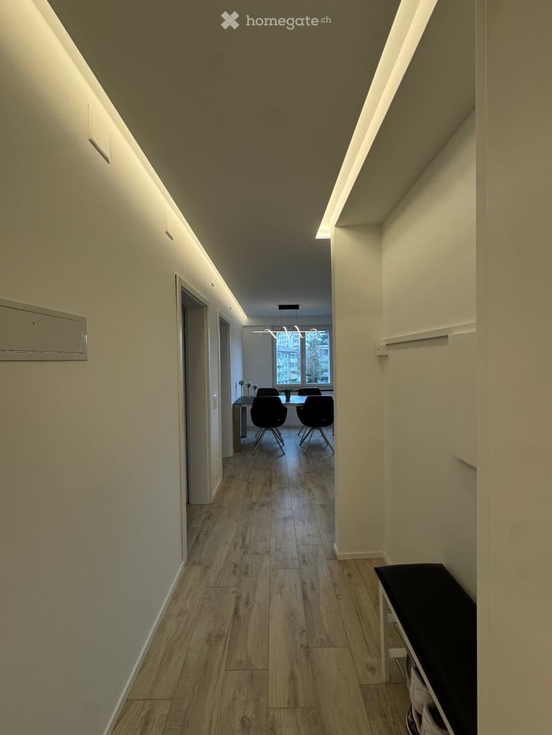 A modern hallway with light wooden flooring, white walls, and built-in lighting. A small seating area with chairs and a table is visible at the end of the corridor.