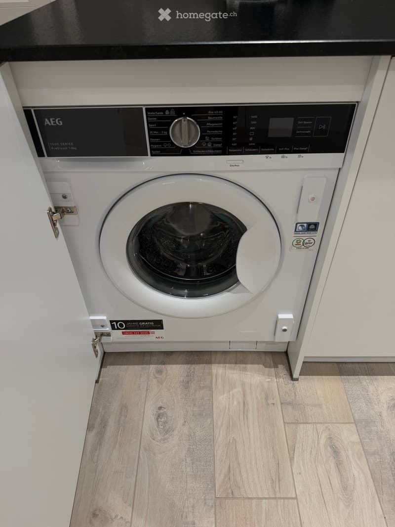 A white AEG washing machine with a control panel is installed in a bathroom with light wooden flooring.