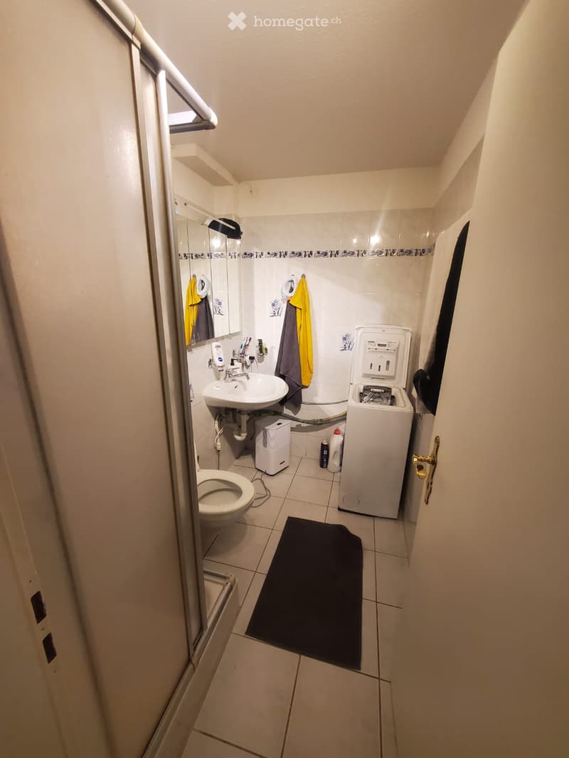Small bathroom with white tiled walls and floor, featuring a sink, toilet, mirror, washing machine, and a black bathroom mat.
