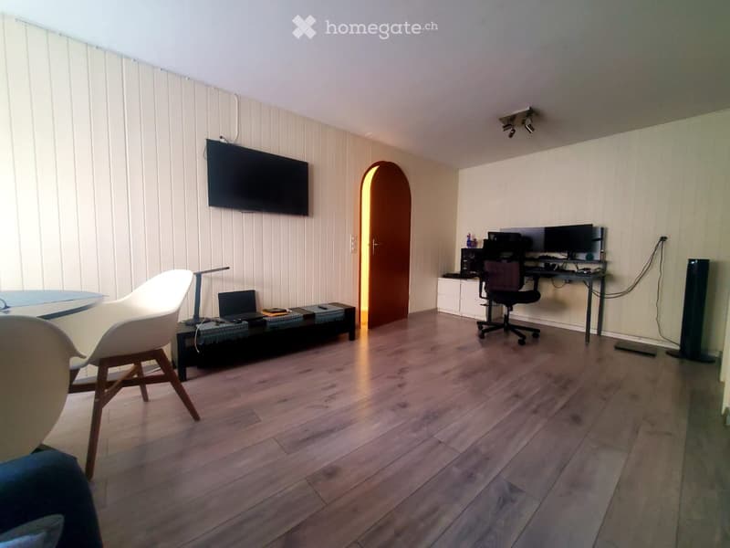 A modern, well-lit room with wooden flooring, a mounted TV, a desk setup with two screens, a comfortable chair, and a small table with a laptop and decorative items.