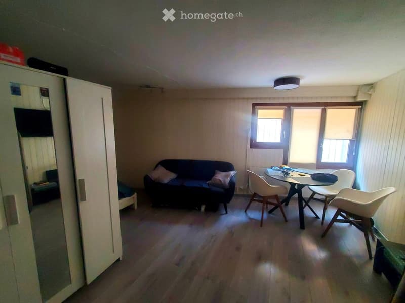 A small, furnished living room with a sofa, a round table with chairs, a mirror, and a bed visible in the background.