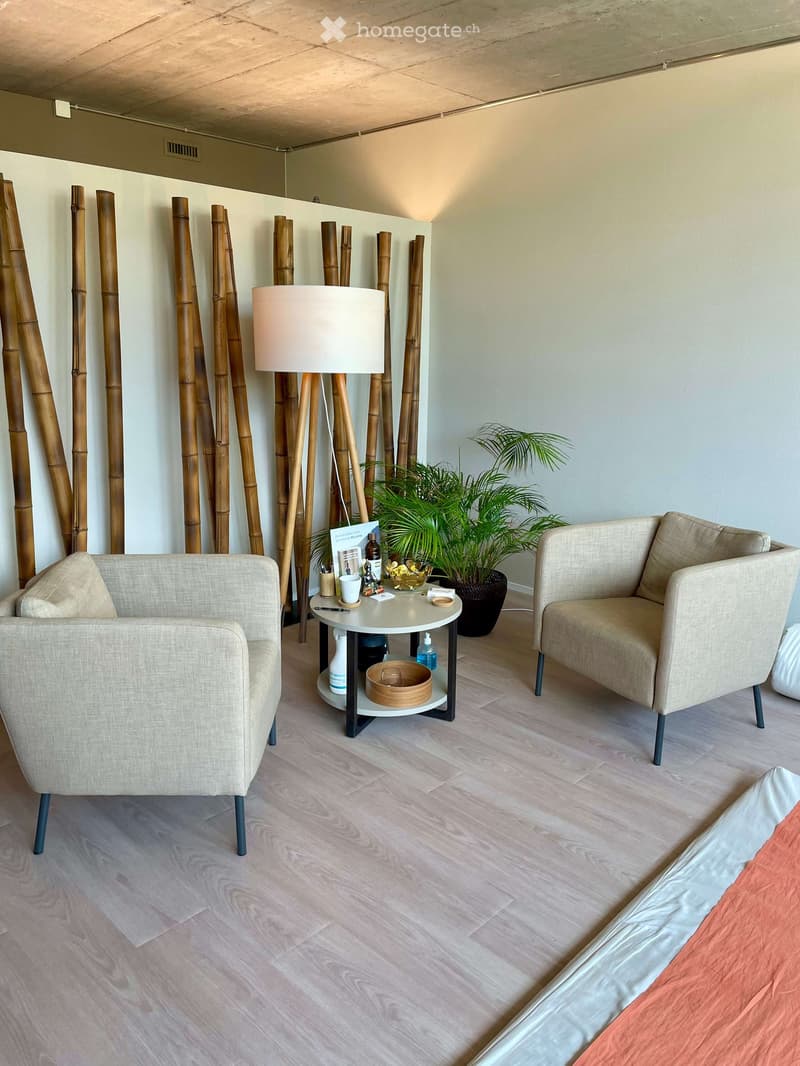 A modern, minimalist living room with light wooden flooring, a beige sofa, a matching armchair, a small round table with a lamp, and a potted plant. Bamboo decorative elements are present on the wall.