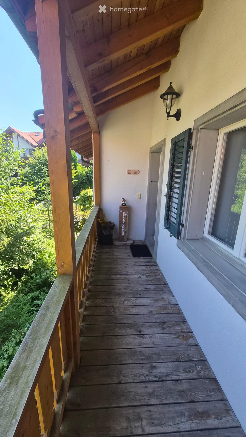 Wooden balcony with railing, steps, and a small welcome sign. The balcony is attached to a building with a light fixture on the wall and a window with shutters.