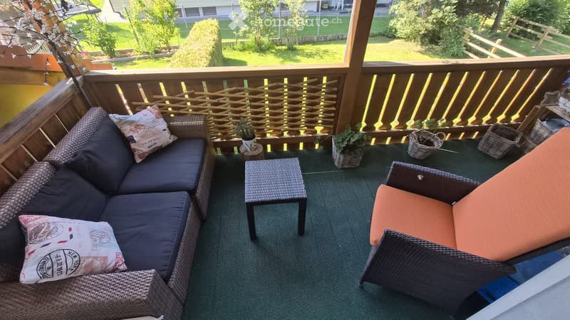 Balcony with wicker furniture, including a sofa, table, and chair, along with potted plants and decorative pillows.