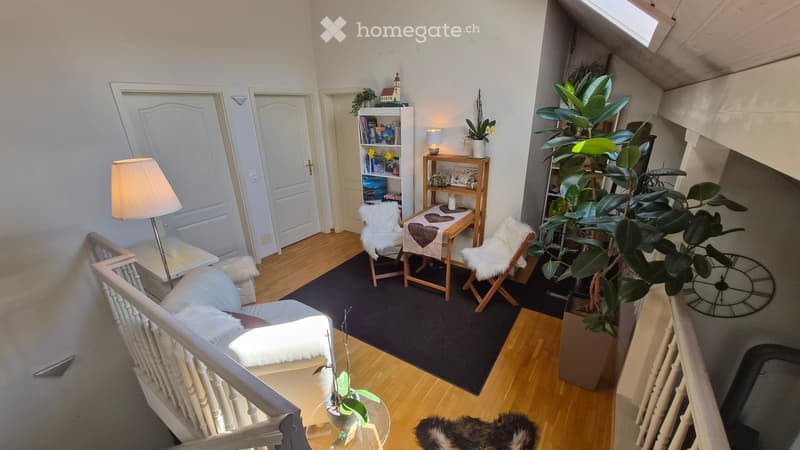 Cozy living room with wooden flooring, white walls, and large windows. Features a white sofa, coffee table, two chairs with fur cushions, a small table with a heart-shaped cushion, a bookshelf, and several plants.