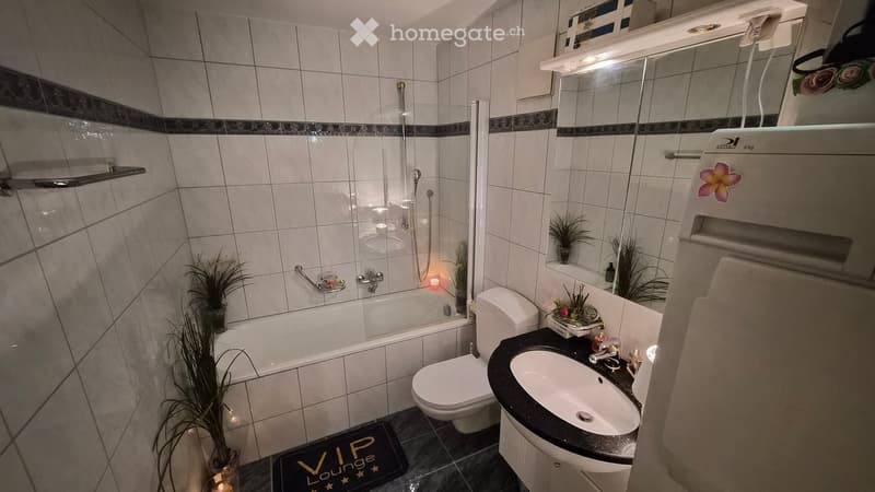 Modern bathroom with a bathtub, toilet, sink, and mirror. Features tiled walls, a glass shower enclosure, and decorative plants.