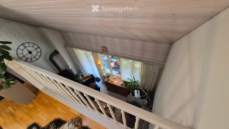 A view from a staircase looking down into a living area with a fireplace, a sofa, a wooden cabinet, and a window with curtains. The ceiling is wooden, and the walls are painted white.