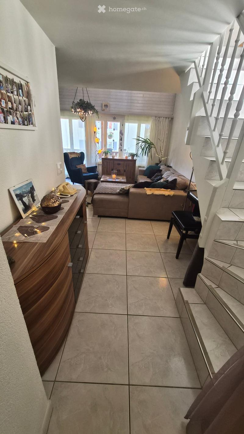 Living room with beige tiled floor, white walls, and a staircase leading upwards. Features a beige sectional sofa, coffee table, wooden cabinet, and a blue armchair.