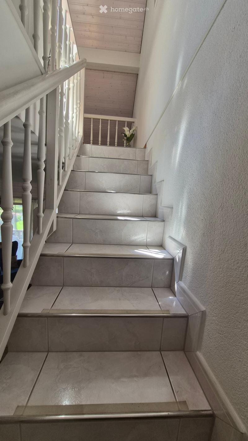 Staircase with white wooden handrail and grey tiled steps, leading to an upper level with a small landing area.