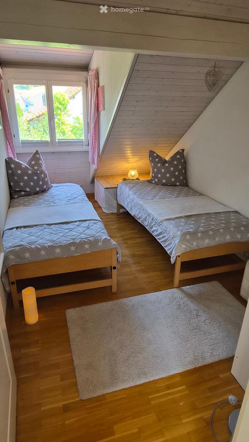 Cozy attic bedroom with two single beds, wooden floors, a nightstand with a lamp, a fluffy rug, and a large window with pink curtains.
