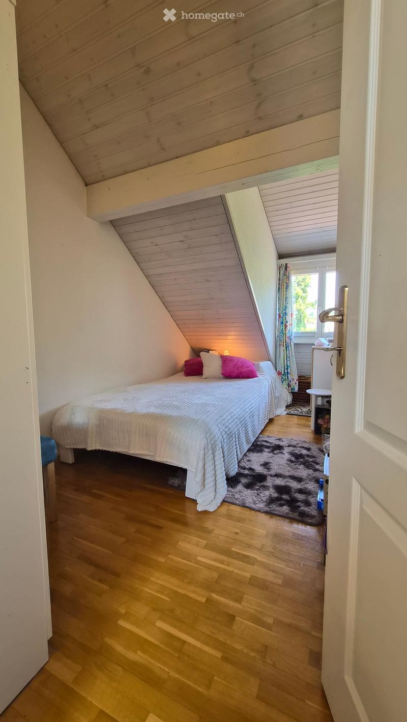 Cozy attic bedroom with wooden floors, slanted wooden ceiling, bed with white bedding and colorful pillows, a small rug, and a window with curtains.
