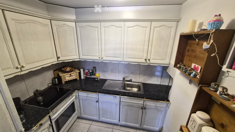 A small kitchen with white cabinets, a black countertop, a sink with a faucet, an oven, and various small decorative items on shelves and the countertop.