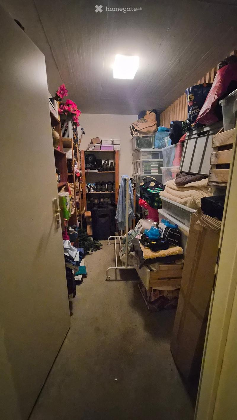 A narrow, cluttered storage area with shelves on both sides filled with various items such as bags, boxes, shoes, and miscellaneous objects. The space has a concrete floor and exposed ceiling beams.