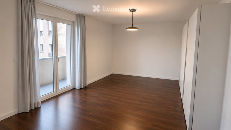 Empty room with wooden flooring, white walls, large sliding glass door with curtains, and a hanging light fixture.