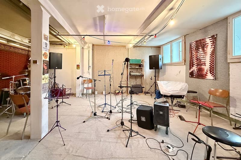 A music studio with a drum set, multiple microphone stands, speakers, a keyboard, and chairs arranged for recording or practice sessions.