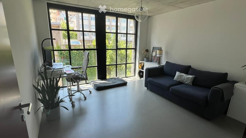Modern living room with large windows, a dark blue sofa, a white desk with a computer, a chair, and a potted plant.