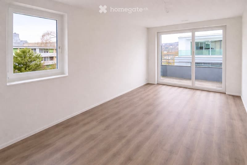 Empty room with light wooden flooring, white walls, large windows, and a sliding glass door leading to a balcony.