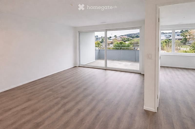 Empty room with light wooden flooring, white walls, large sliding glass doors leading to a balcony, and a window with a view of the neighborhood.