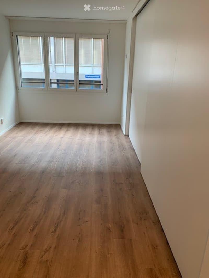 Empty room with wooden flooring and white walls, featuring a large window with blinds and a view of a building outside.
