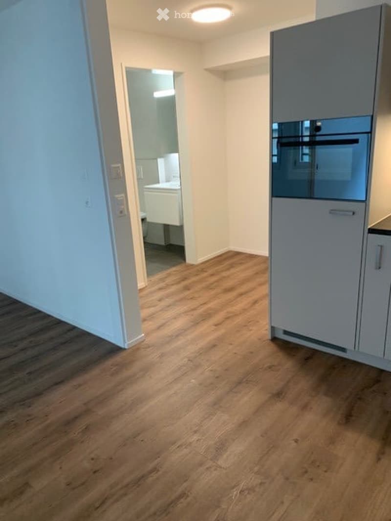 Empty room with wooden flooring, white walls, and a partially visible kitchen area with cabinets and a sink in the background.