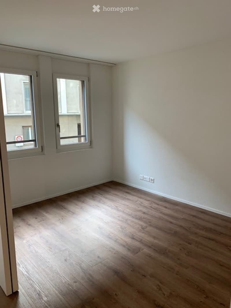 Empty room with wooden flooring, white walls, and two windows allowing natural light.