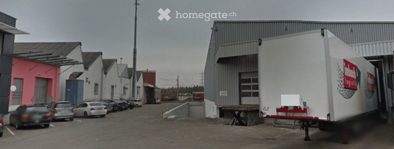 Industrial building with a large garage entrance, several parked cars, and a logistics trailer nearby.