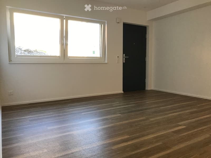 Empty room with wooden flooring, white walls, large window, and a black door.