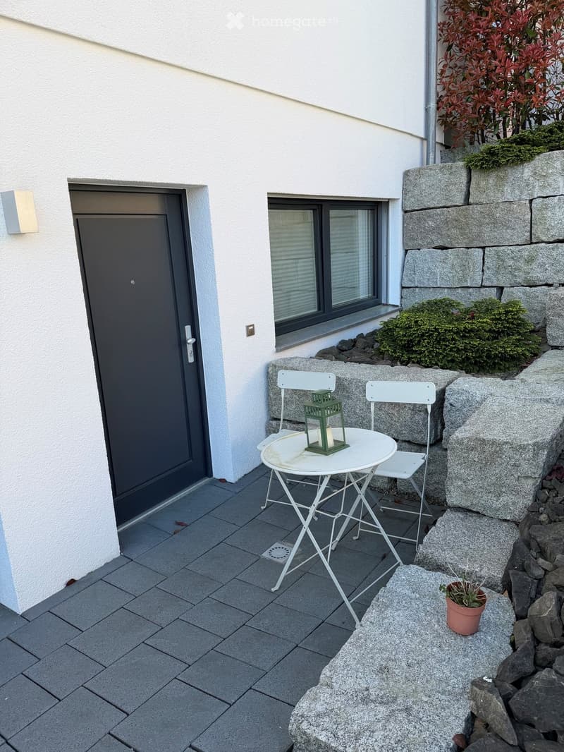 Small outdoor seating area with a white table, two white chairs, a green lantern, and a potted plant on a stone ledge.