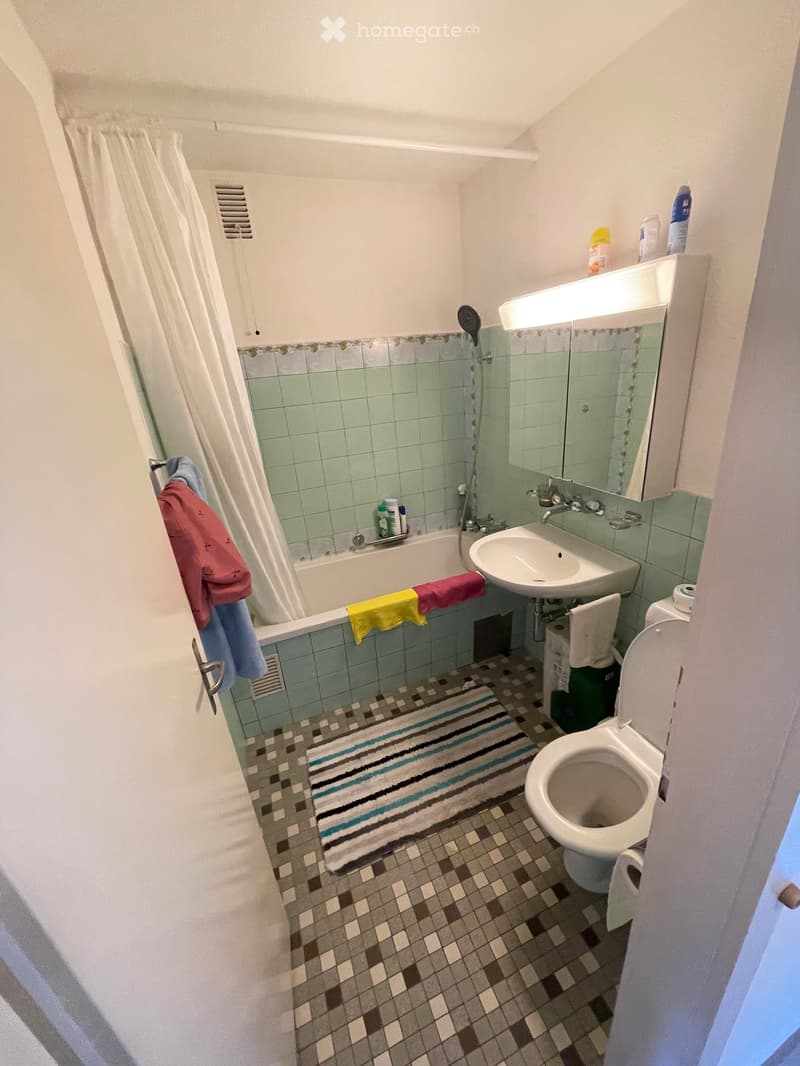 Bathroom with a bathtub, sink, toilet, mirror, and various towels. The walls are tiled in a light green color, and the floor has a patterned tile design.