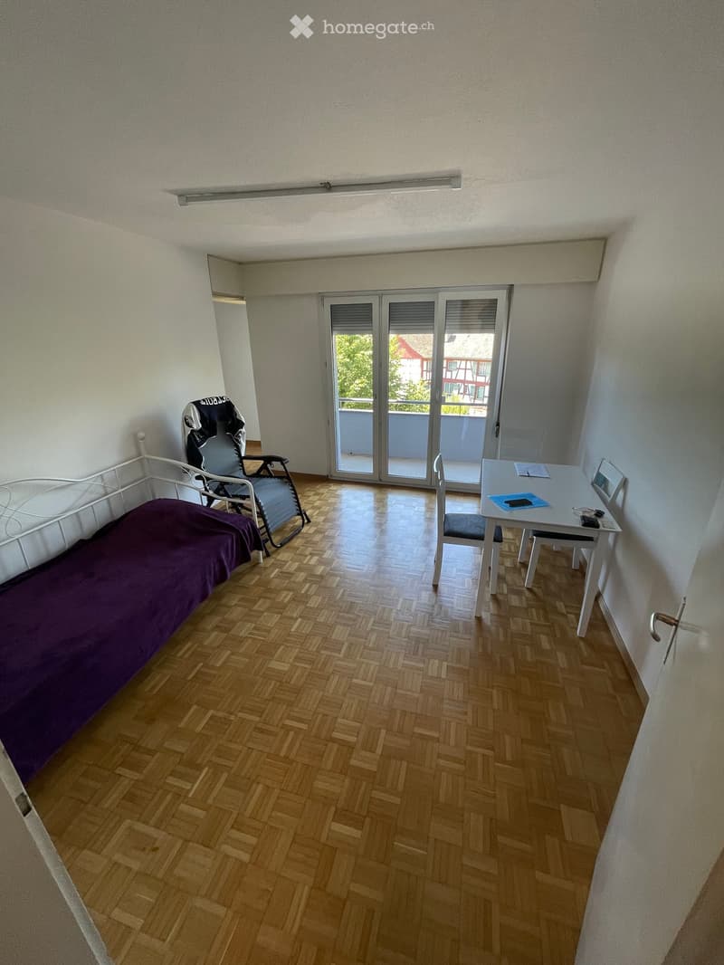 A furnished room with a bed, a chair, a small table with a laptop, and a sliding glass door leading to a balcony.