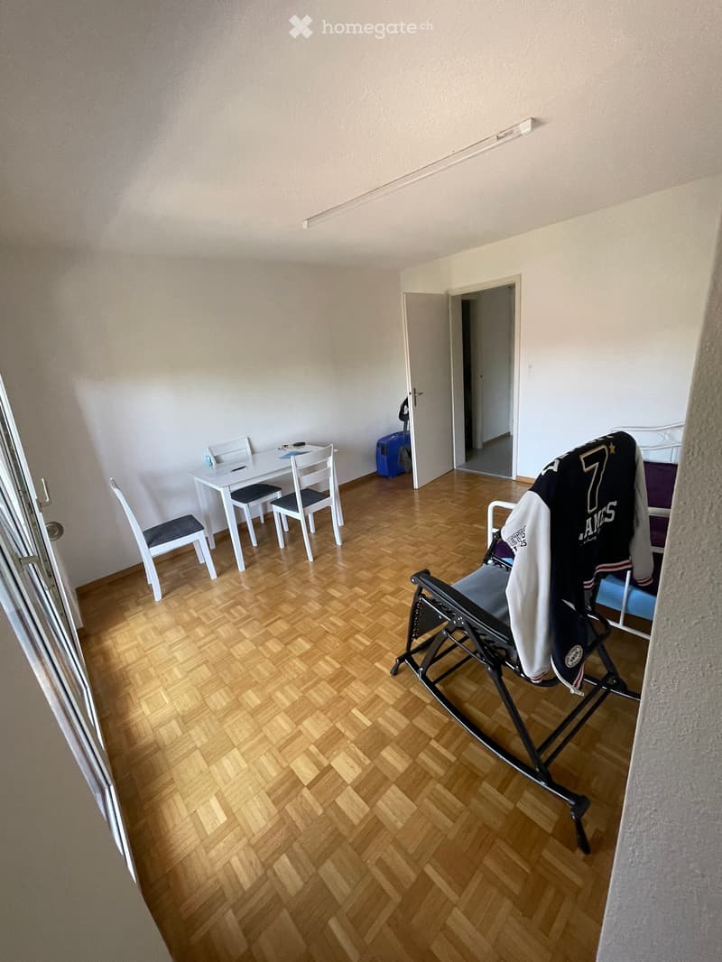 A furnished room with a wooden floor, a small white table with chairs, a jacket draped over a chair, and a partially visible bed in the background.