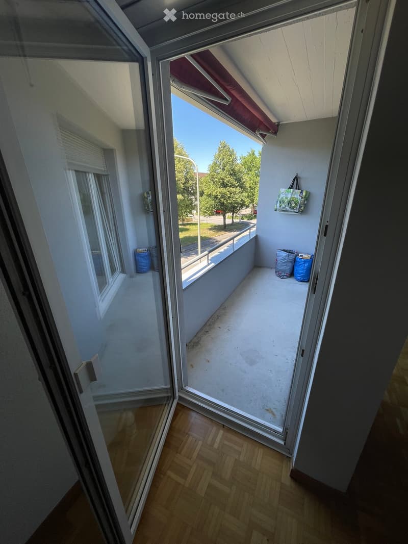 Balcony with wooden floor, white walls, and a view of the street and trees. There are a few bags and a hanging bag on the wall.