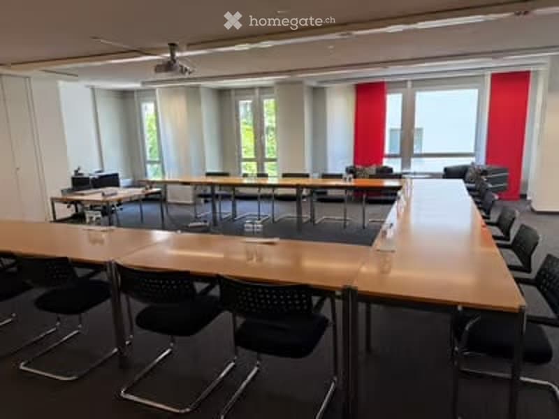 Conference room with long wooden table, black chairs, projector, and screen. Large windows with natural light.
