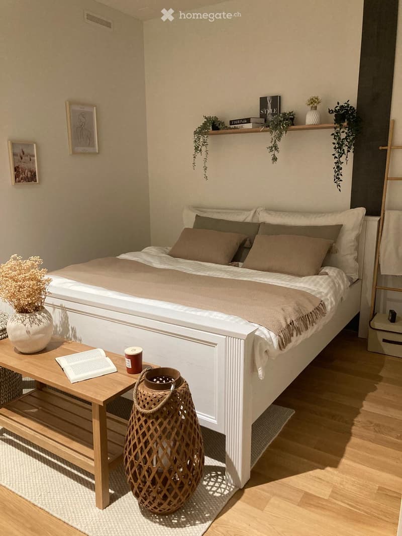 A modern, well-lit bedroom with a large bed, nightstand, and decorative items including a vase with dried flowers, a book, and a cup on a small table. The room features wooden flooring and a cozy, minimalist design.