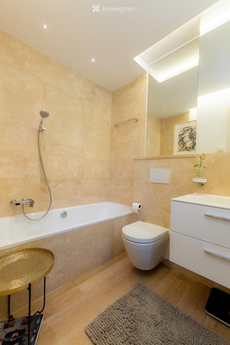 Modern bathroom with beige tiles, a bathtub, toilet, sink, mirror, and a small round table with a flower vase.