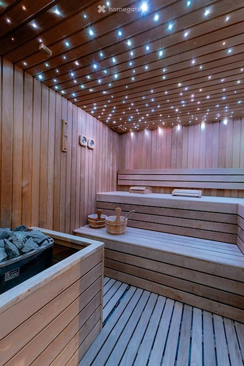 A wooden sauna interior with wooden benches, steps, and a small black container. The ceiling and walls are made of wood, and the ceiling is adorned with small, evenly spaced lights.